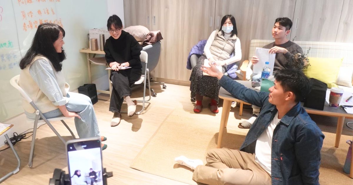 團督演練現場：一位成員坐著演練，數人在旁觀察，有人低頭記筆記，手機架在腳架上錄影